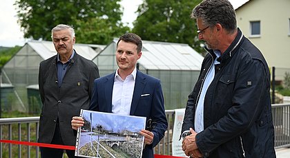 Auf dem Bild sind drei Männer zu sehen. Einer hält ein Foto in der Hand. 