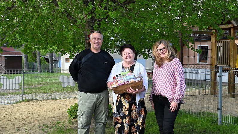 Auf dem Foto sind drei Personen zu sehen. Eine Frau hält einen Präsentkorb vor sich und lächelt. 