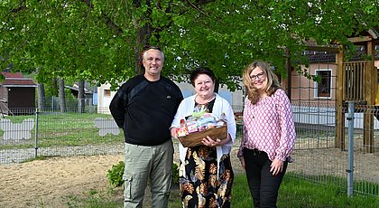 Auf dem Foto sind drei Personen zu sehen. Eine Frau hält einen Präsentkorb vor sich und lächelt. 