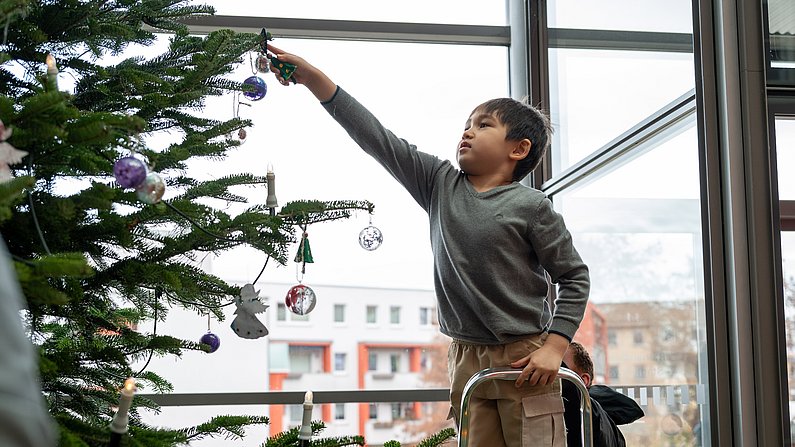 Auf dem Bild ist ein Kind zu sehen, welches auf einer Leiter steht und einen Weihnachtsbaum schmückt.