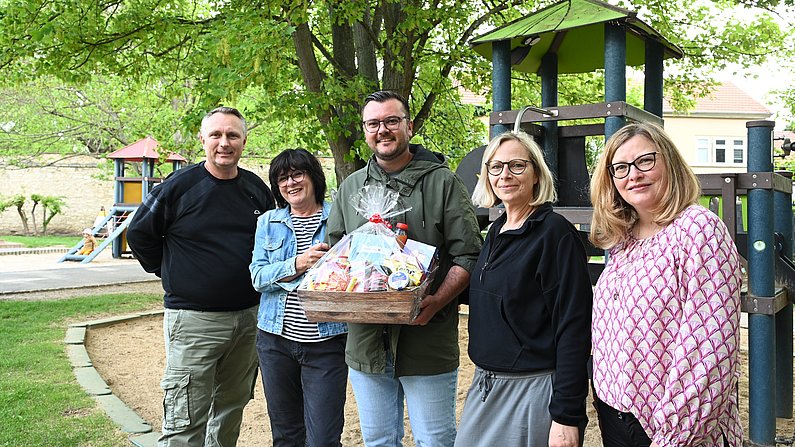 Auf dem Foto sind fünf Erwachsene zu sehen. Ein Mann hält einen Präsentkorb vor sich. Alle lächeln. 