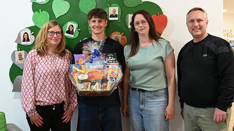 Auf dem Foto sind vier erwachsene Personen zu sehen. Ein Mann hält einen Präsentkorb vor sich. Alle wirken fröhlich. 