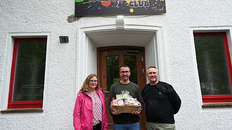 Auf dem Foto sind drei Personen zu sehen. Ein Mann hält einen Präsentkorb vor sich und lächelt