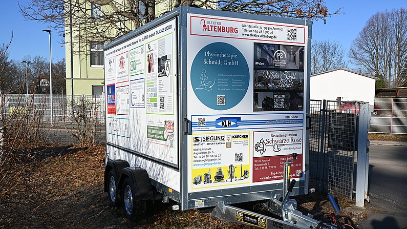 Auf dem Foto ist eine Fahrzeuganhäger zu sehen. Auf der Plane des Anhängers sind viele Firmenlogos zu sehen. 