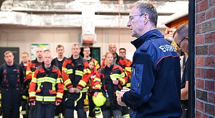 Auf dem Foto sieht man einen Mann der zu einer Gruppe Feuerwehrleuten spricht. Er trägt eine blaue Jacke mit Feuerwehraufschrift. 