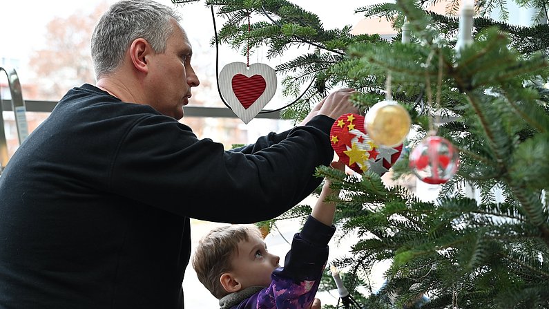 Auf dem Foto ist ein Mann und ein Kind zu sehen, beide schmücken einen Weihnachtsbaum. 