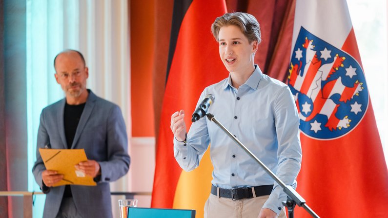 Auf dem Bild sind zwei Personen zu sehen. Die eine Person spricht am Mikrofon, die andere steht hinter ihm. Auf dem Bild sind zwei Personen zu sehen. Die eine Person spricht am Mikrofon, die andere steht hinter ihm.