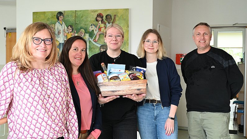 Auf dem Foto sind fünf erwachsene Personen zu sehen. Eine Frau hält einen Präsentkorb vor sich. Alle lächeln. 