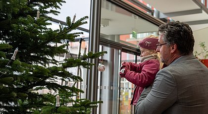 Auf dem Foto ist ein mann zu sehen, der ein Kind hoch hlt, dass es an einen Weihnachtsbaum ein Dekoelement hängen kann. 