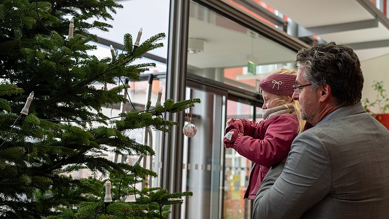 Auf dem Foto ist ein mann zu sehen, der ein Kind hoch hlt, dass es an einen Weihnachtsbaum ein Dekoelement hängen kann. 