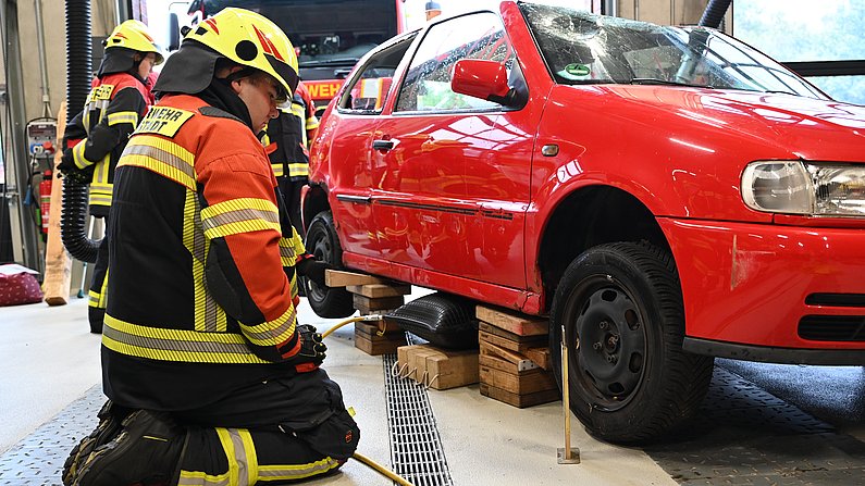 Auf dem Foto sieht man einen Mann, der vor einem roten Auto kniet. Das Auto ist aufgebockt um Räder zu wechseln. 