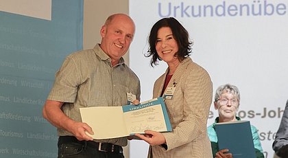 Auf dem Foto ist ein Mann und eine Frau zu sehen. Beide halten eine Urkunde in den Händen und lächeln in die Kamera. 